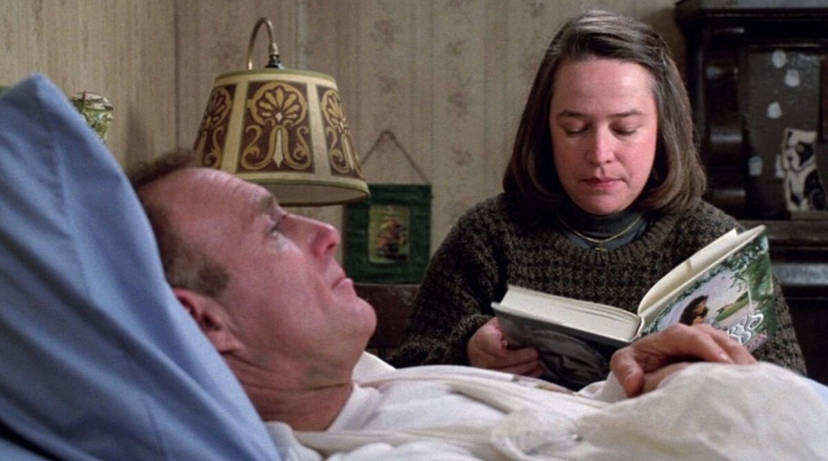 Homem deitado numa cama com a cabeça apoiada em um travesseiro. Ele está com o braço enfaixado. À sua frente, uma mulher sentada na cabeça da cama, lendo um livro enquanto usa um agasalho. Cena de "Louca Obsessão", do Stephen King.