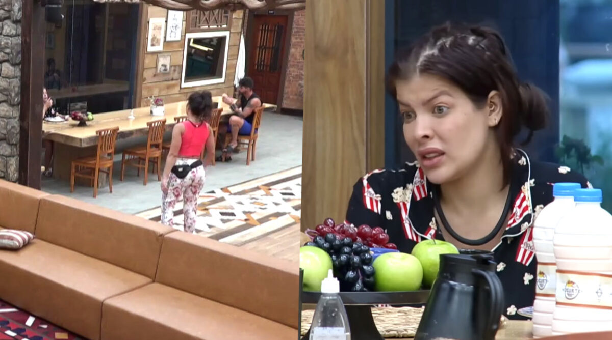 Mulher usando camiseta rosa e calça leg branca em pé em frente a uma mesa de cozinha, onde outra mulher de pijama está sentada olhando fixamente pra ela. As duas são Nadja e Jaquelline, participantes de A Fazenda 15, respectivamente.