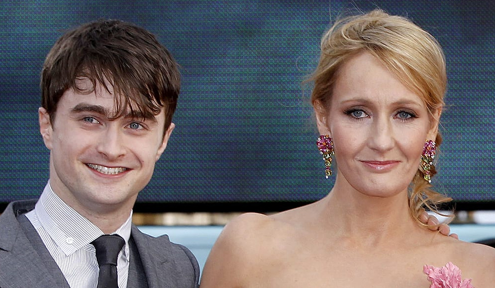 Daniel Radcliffe usando terno e gravata e J.K. Rowling com cabelo preso e sorrindo para a câmera.